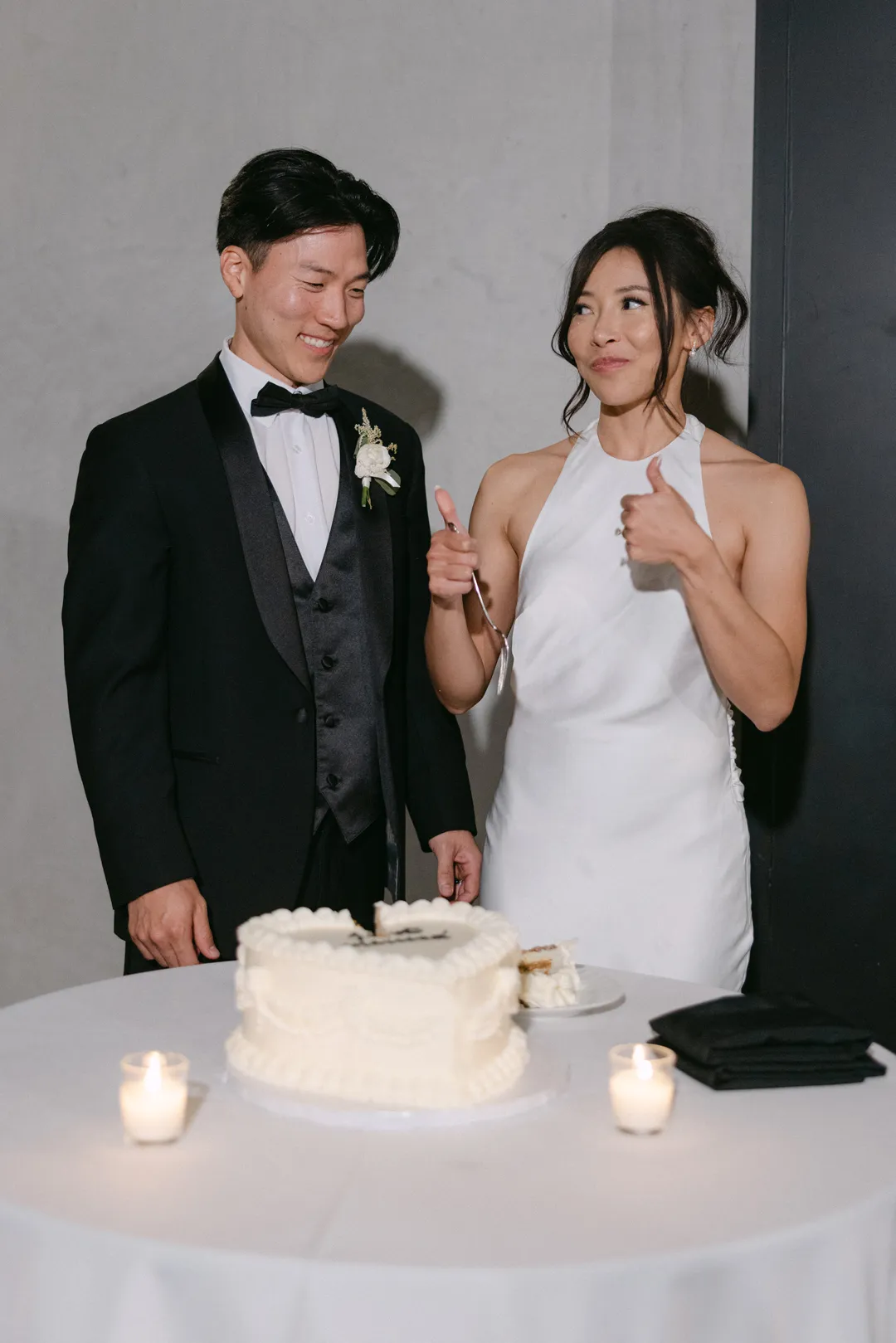 Newlyweds smiling and giving thumbs-up beside the cake.