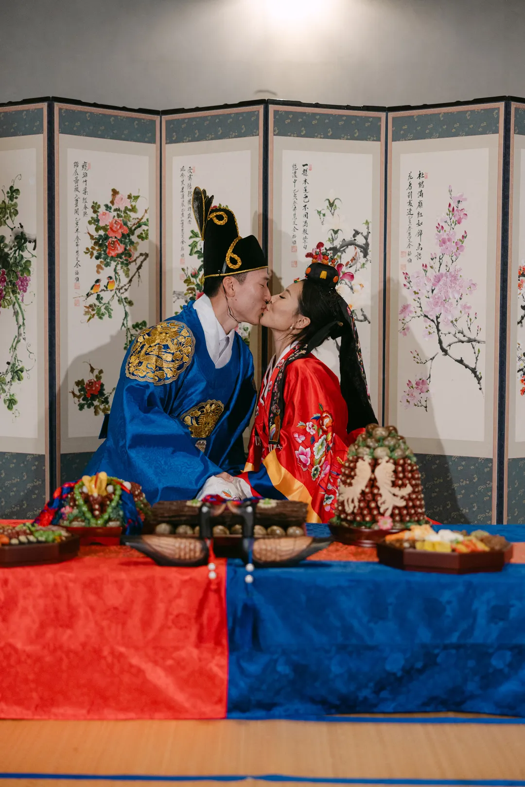Wedding couple in red and blue ceremonial clothing kissing beside a floral folding screen.