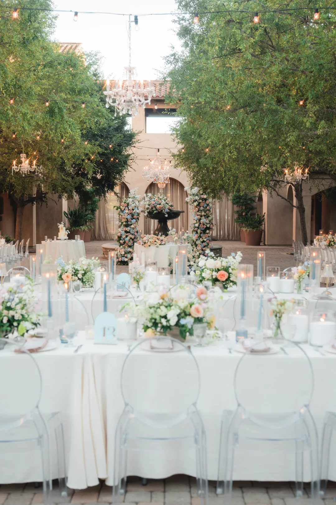 White-linen reception tables with clear chairs and pastel flowers.