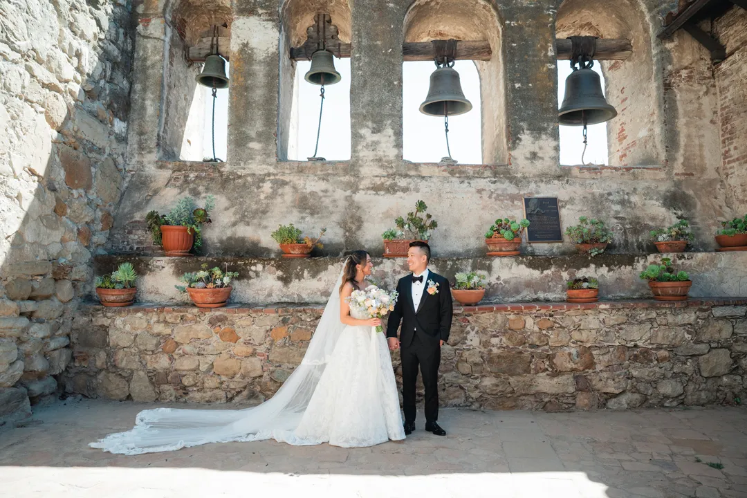 Wedding couple posing beneath four bells at Mission San Juan Capistrano.