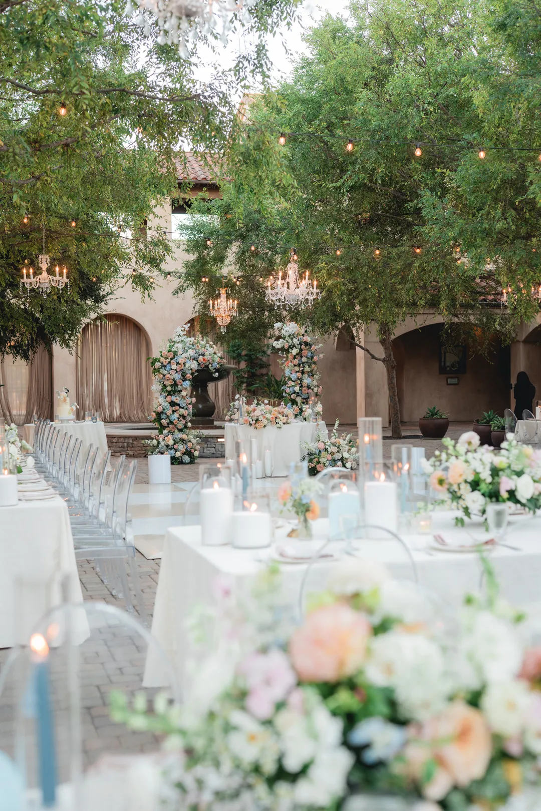 Serra Plaza reception table beneath trees and chandeliers, with blue candles and blush napkins.