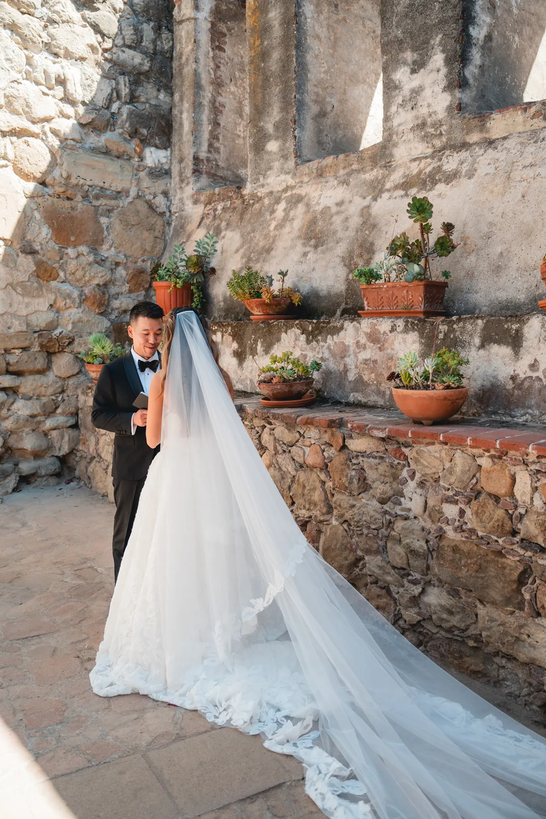 Wedding couple embracing beneath the Mission San Juan Capistrano bells, with a long veil trailing behind.