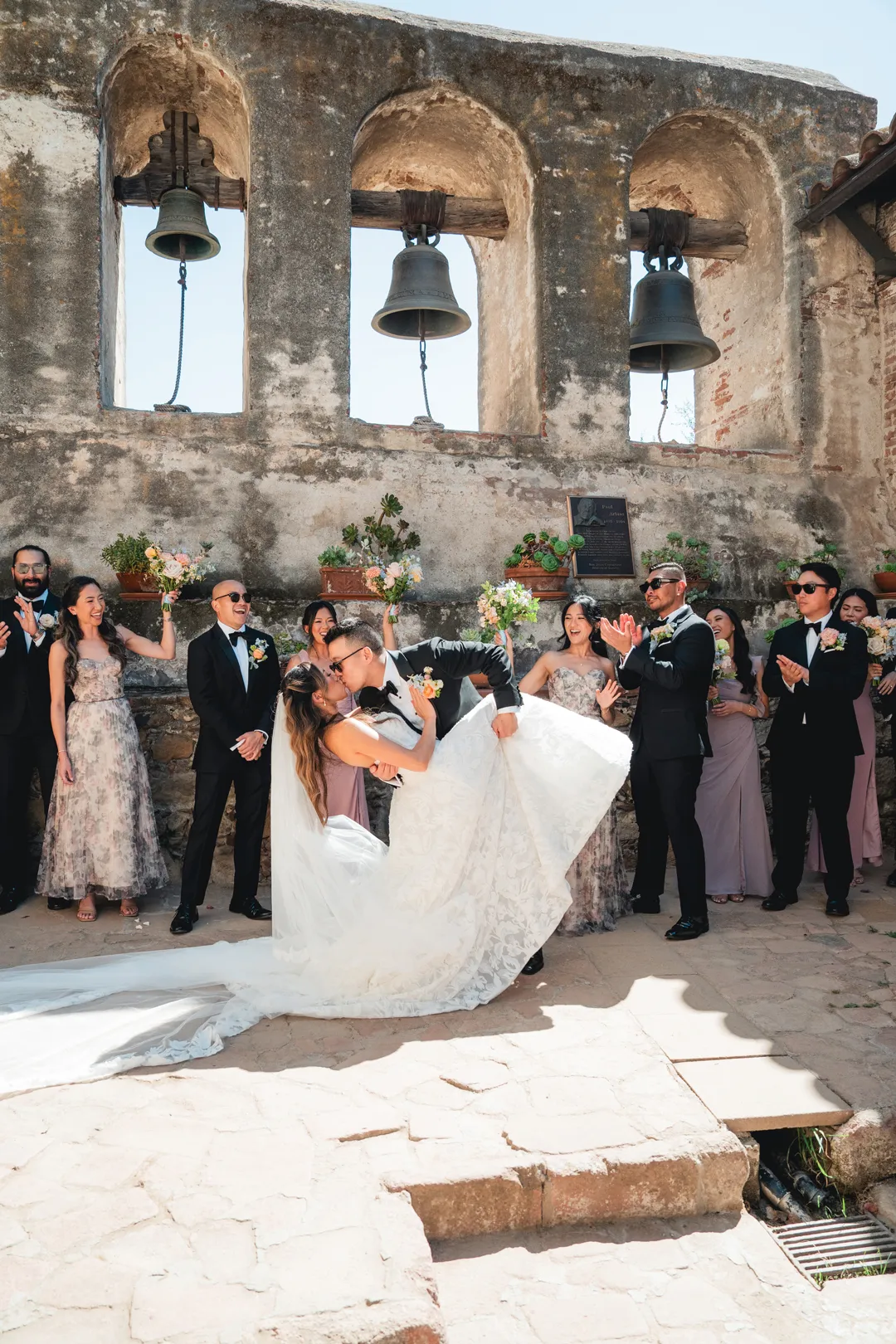 Groom dipping the bride as the wedding party cheers beneath the Mission San Juan Capistrano bells.