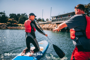 A person wearing a life jacket kneels on a paddleboard while an instructor gestures nearby.