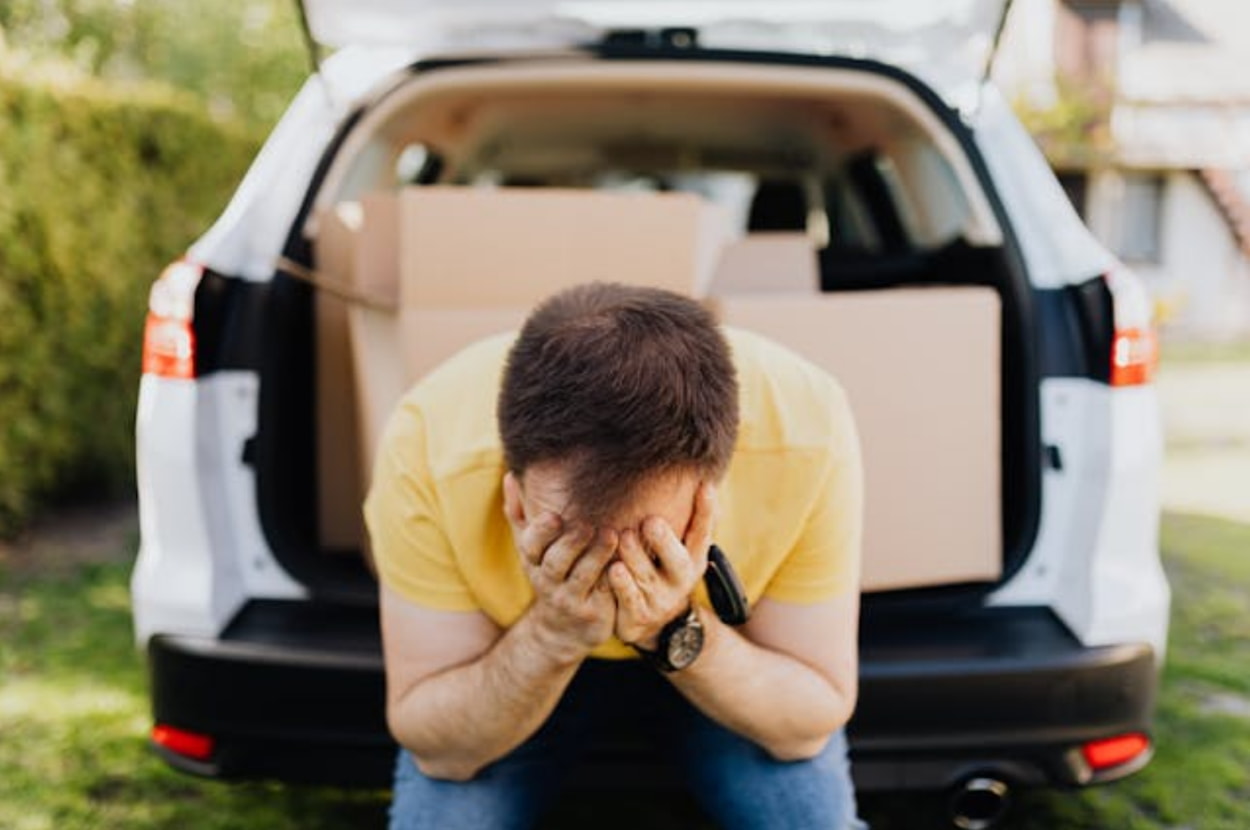 A distressed man sits in the trunk of a car, with his head in his hands, reflecting a moment of despair. 
