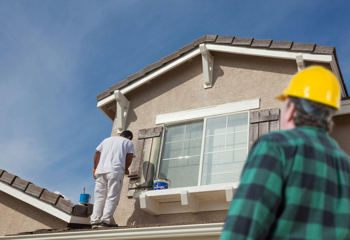 Two men using a ladder to paint the exterior of a house, focused on their task under a clear blue sky.