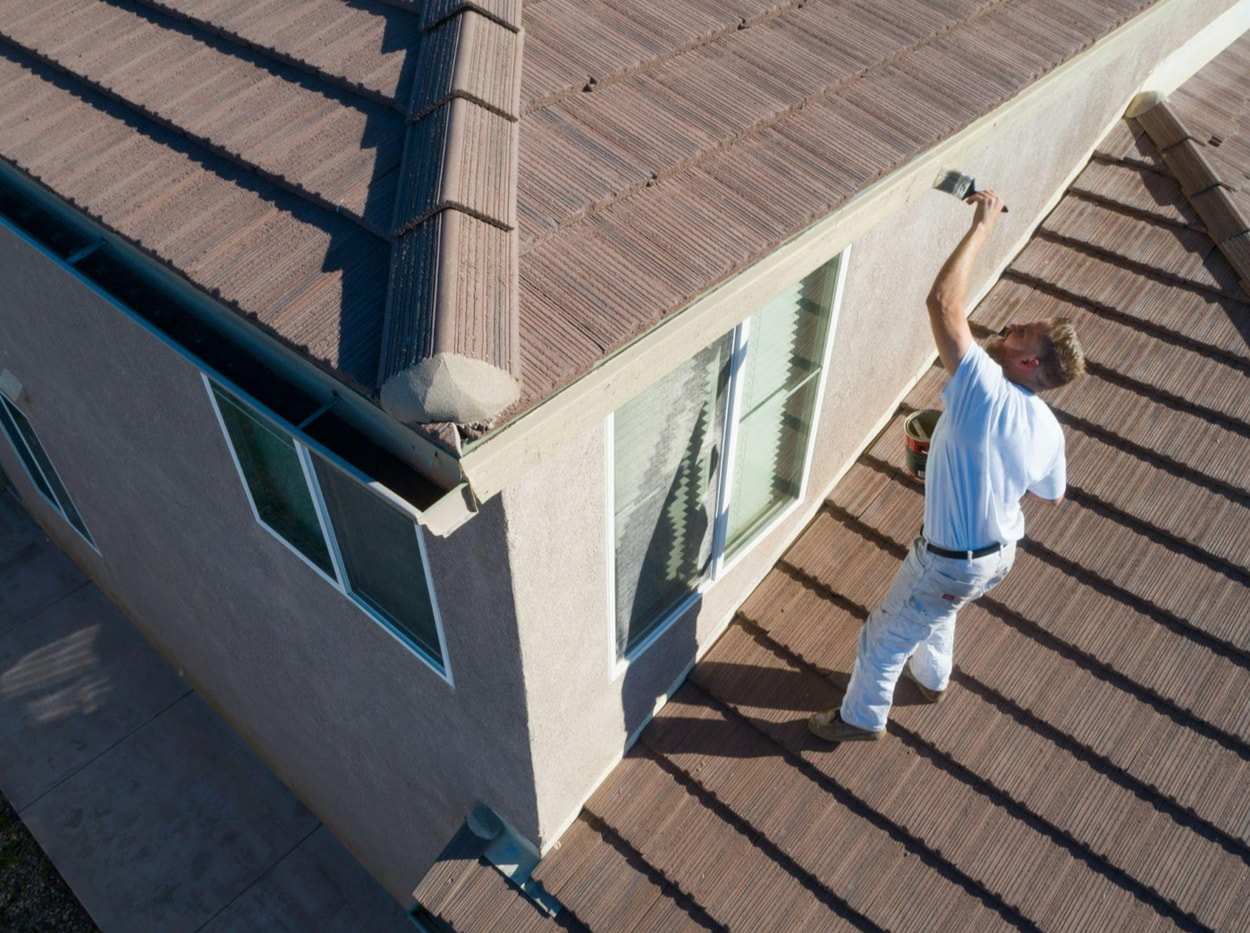 A man applying paint to the roof of a house using a brush while standing on a ladder.