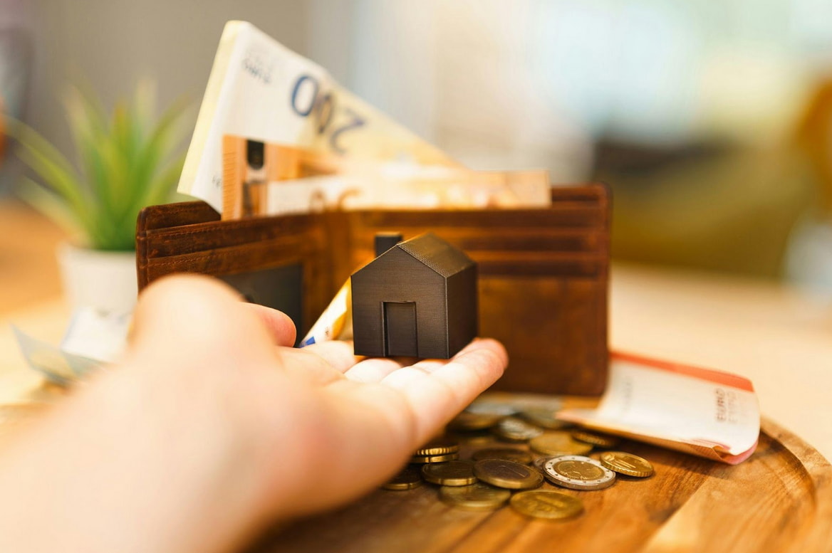 A hand holds a wallet filled with cash next to a small model house, symbolizing financial security and home ownership.