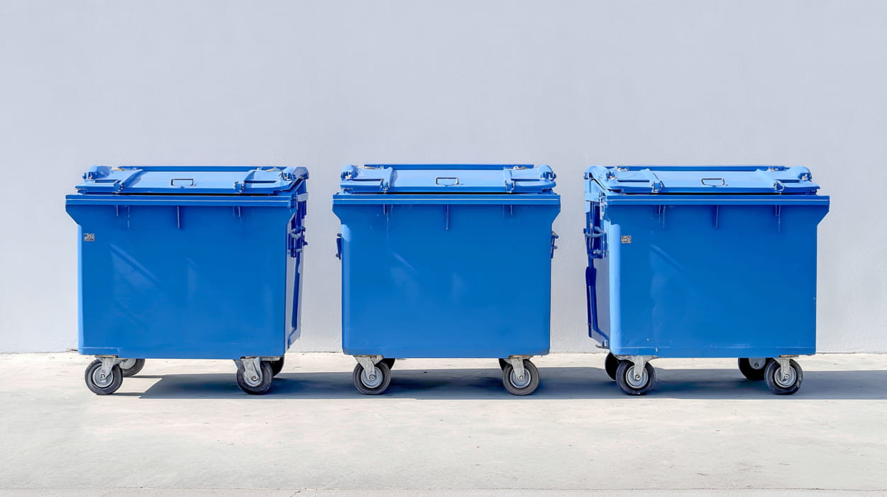 Three blue wheeled garbage containers positioned in front of a plain white wall.