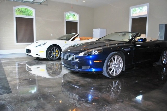 Two luxury convertibles, a white and a dark blue car, are parked in a pristine garage with glossy floors. Natural light streams in from the windows.