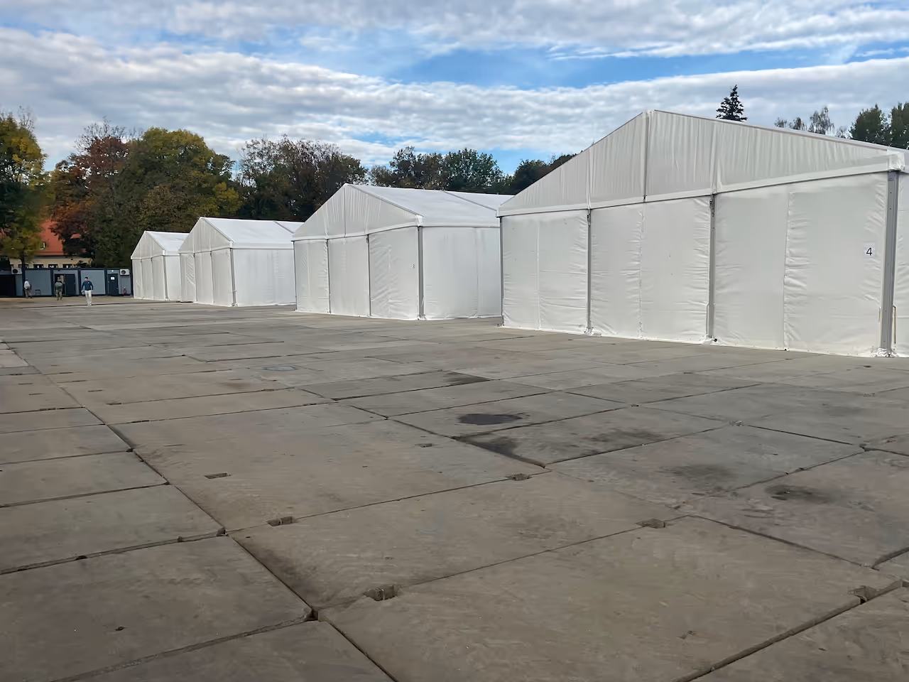 Row of large white temporary tents on a paved area with trees and a partly cloudy sky in the background.