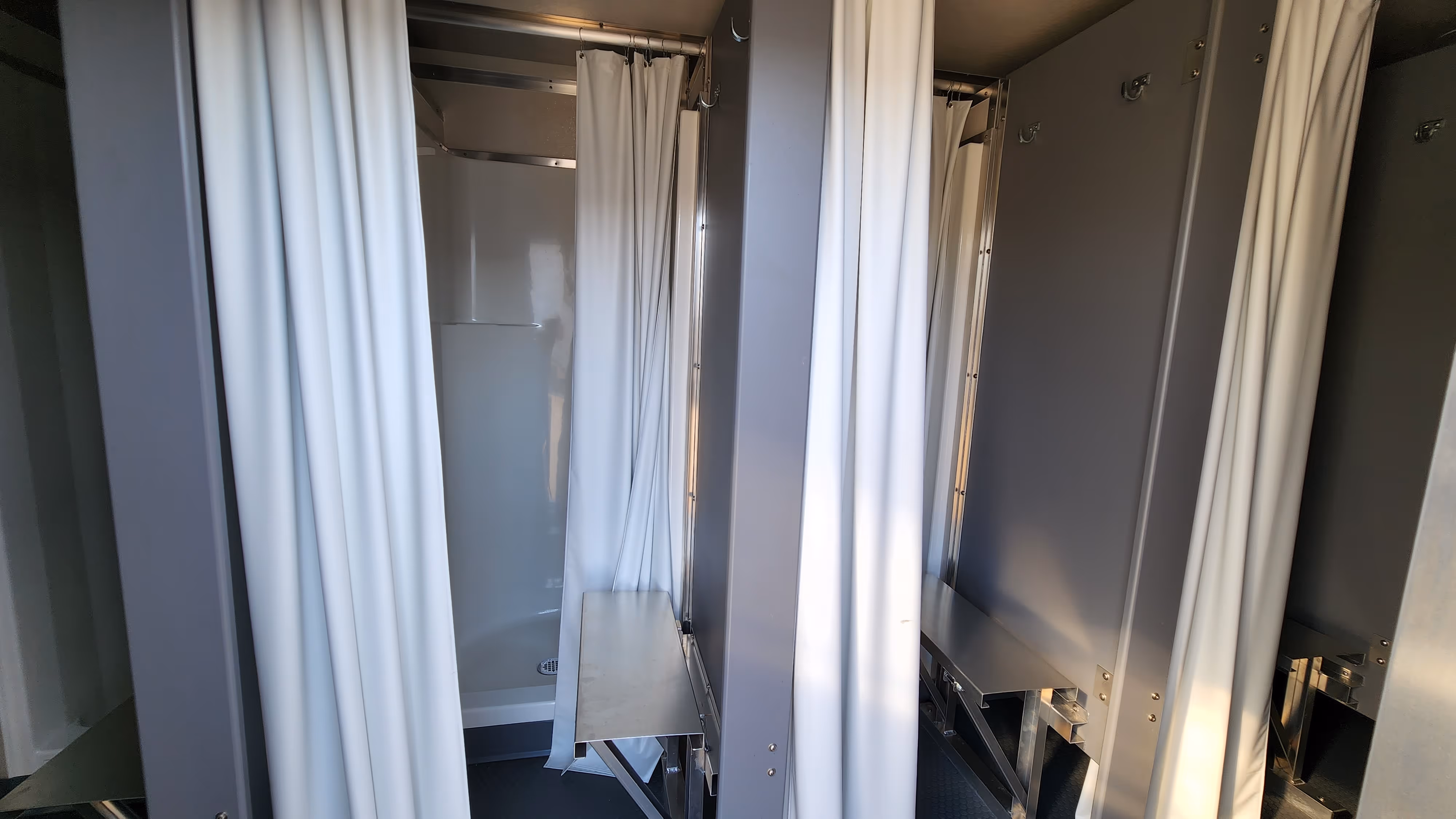 Two shower stalls with white curtains and fold-down metal seats inside.