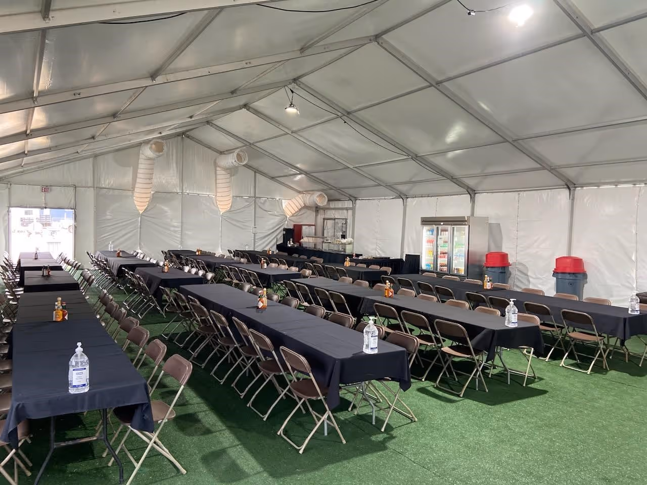 Interior of a large event tent with rows of tables covered in black tablecloths and brown folding chairs, hand sanitizer and condiment bottles on tables.
