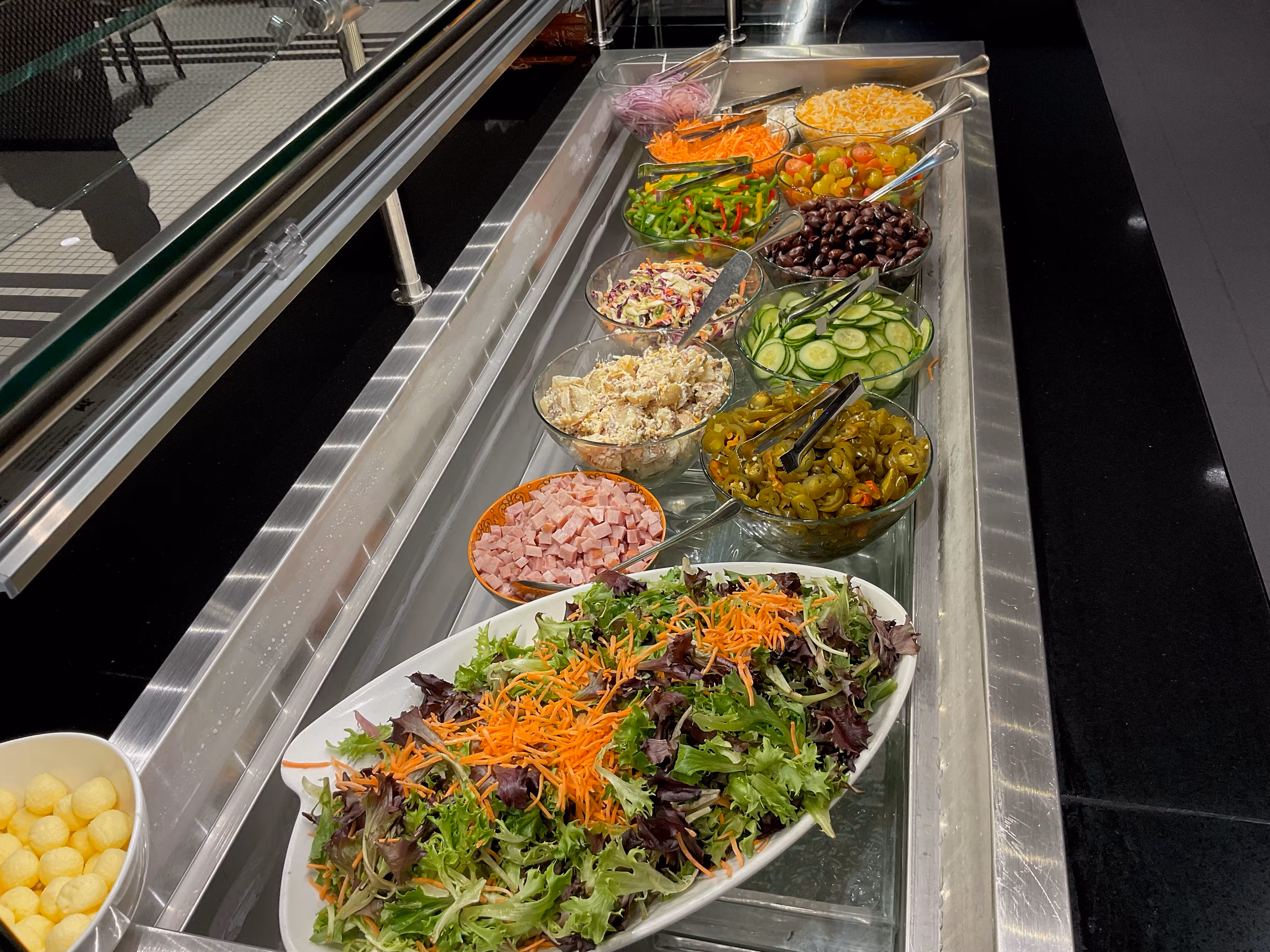 Salad bar with bowls of mixed greens, shredded carrots, diced ham, jalapeños, cucumbers, olives, cherry tomatoes, shredded cheese, and coleslaw.