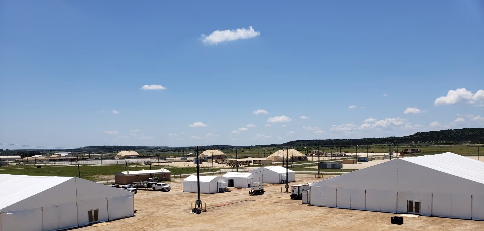 Open outdoor area with large white tents, utility trucks, and green fields under a clear blue sky with scattered clouds.
