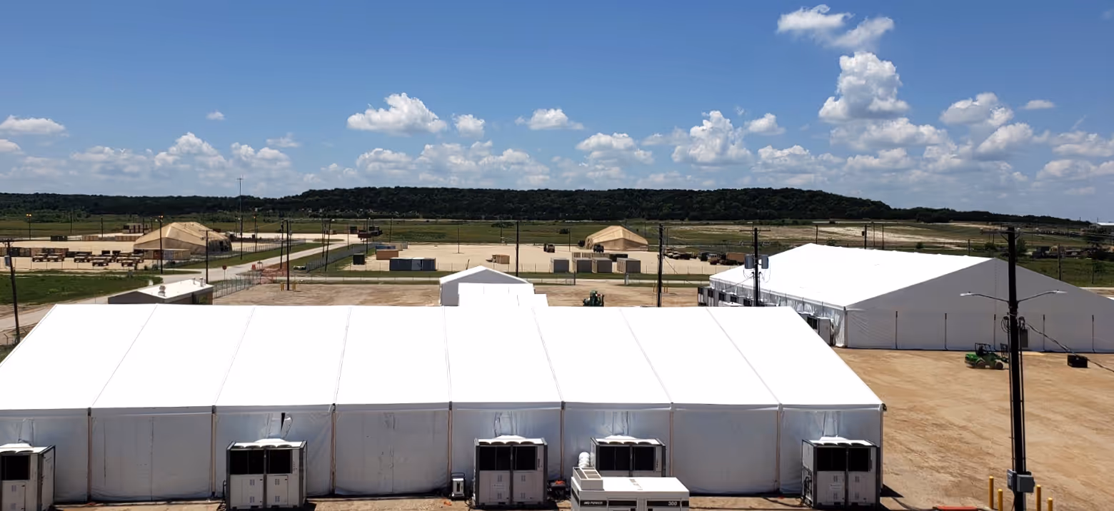 Large white temporary canopy tents set up on a sandy area under a blue sky with scattered clouds.