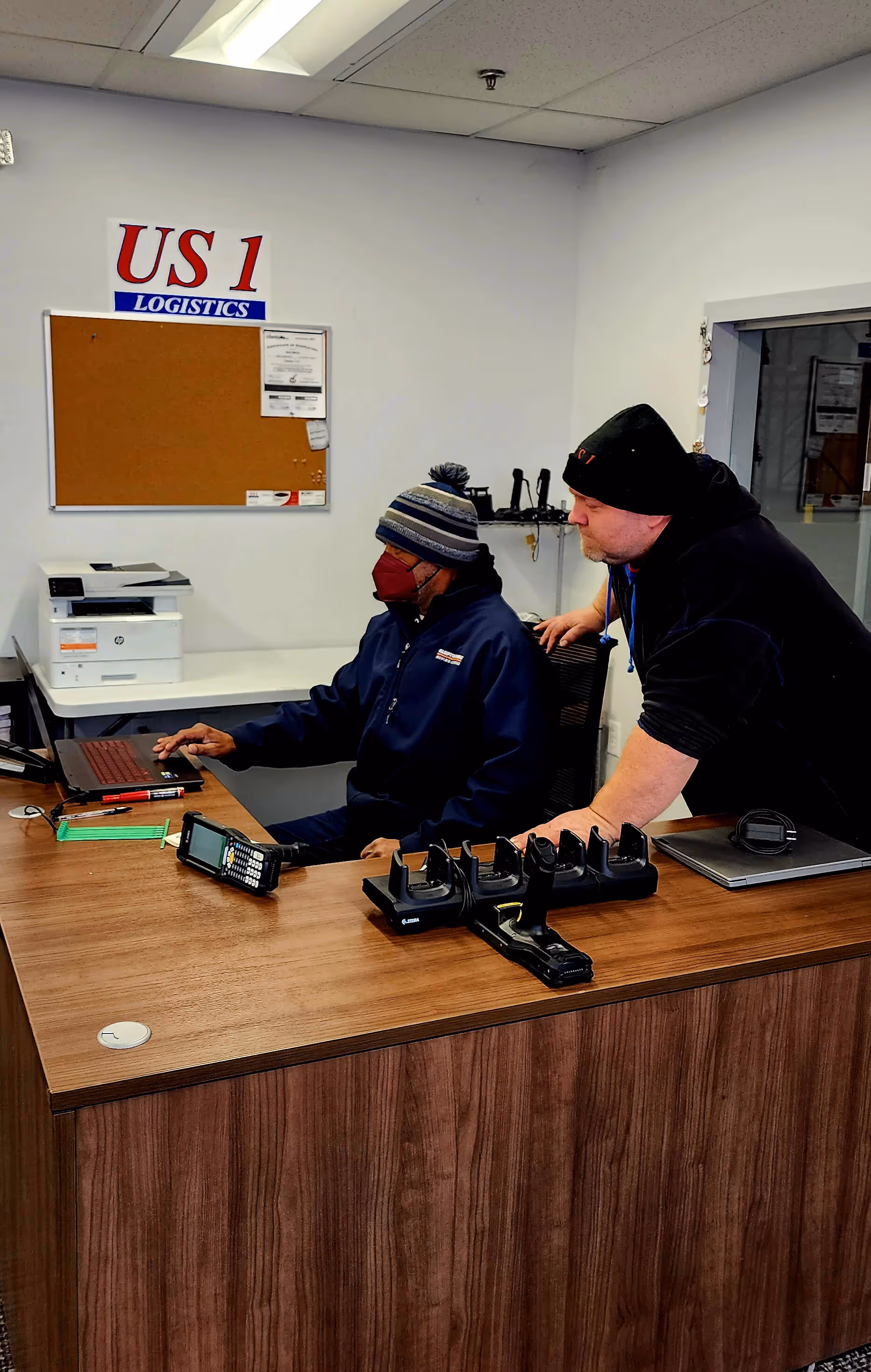 Two men in winter hats working at a desk with a laptop, barcode scanner, and US 1 Logistics sign behind them.