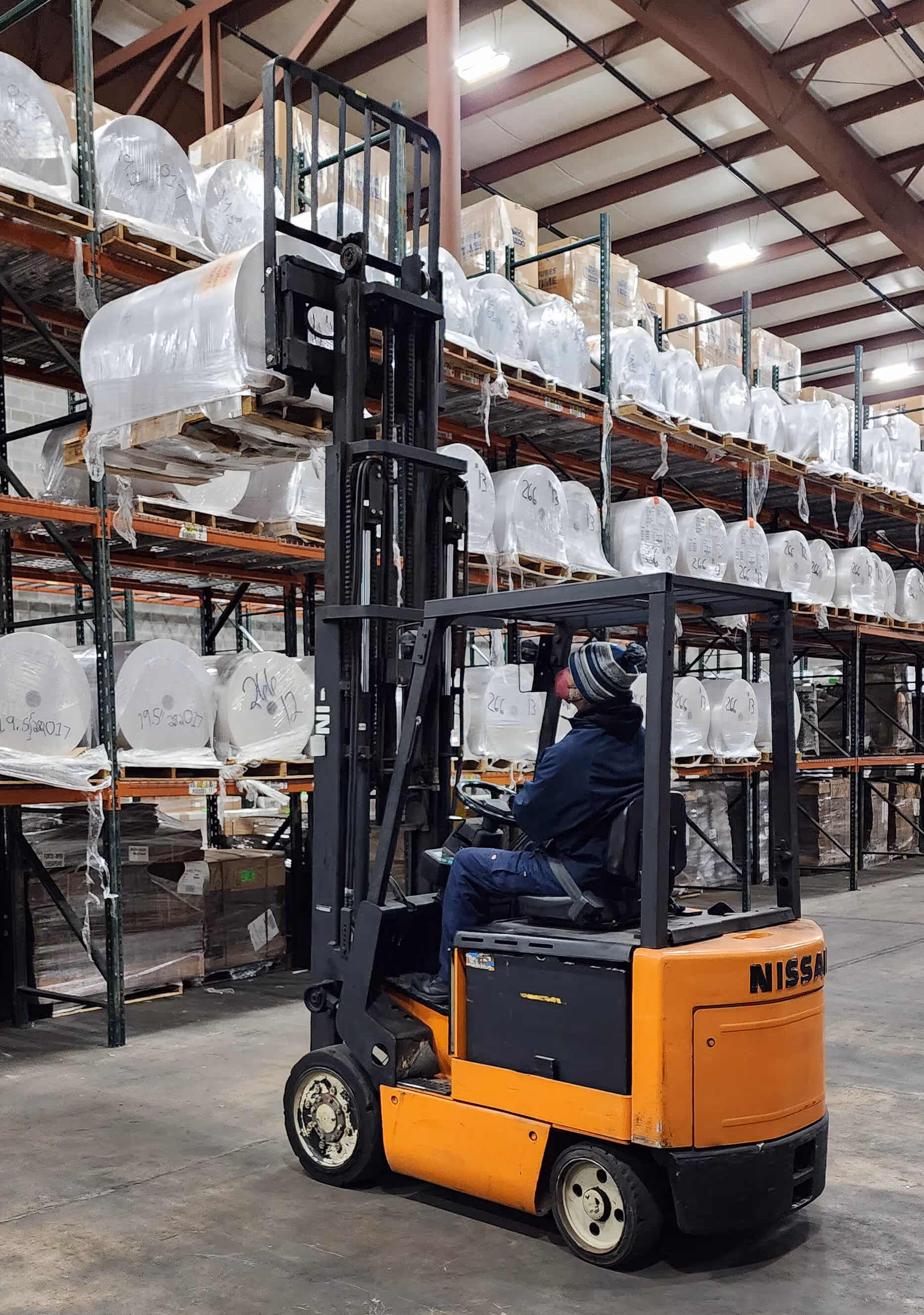 Worker wearing a hat and mask operates an orange Nissan forklift in a warehouse with stacked white rolls.