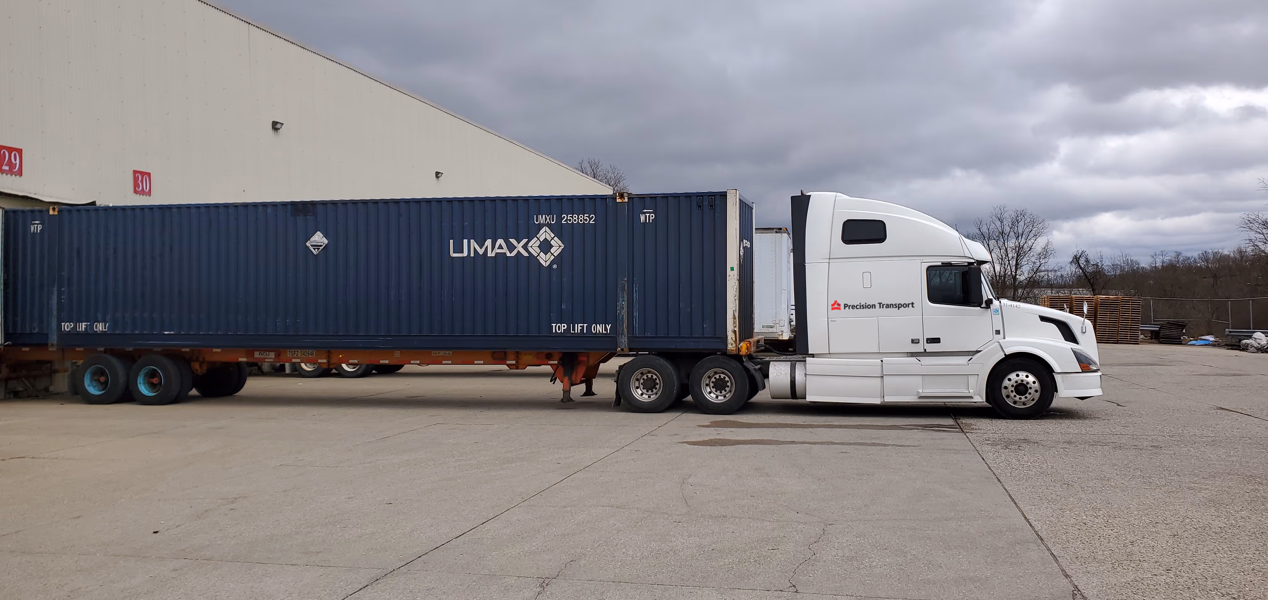 White Precision Transport semi truck attached to blue UMAX shipping container on concrete lot under a cloudy sky.