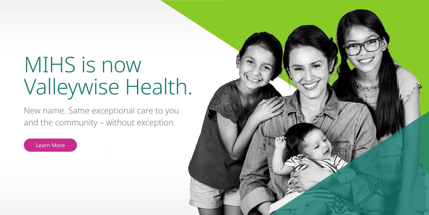 A header on the redesigned Valleywise Health, with a black and white cut out image of a young hispanic mother smiling with her two pre-teen daughters and a her baby in her arms. The image sits between two brand-colored triangles with varying degrees of transparency. The header text reads "MIHS is now Valleywise Health."