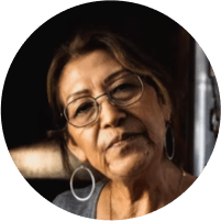 A middle-aged Hispanic woman with brown hair tied up in a ponytail, wearing glasses and earings.