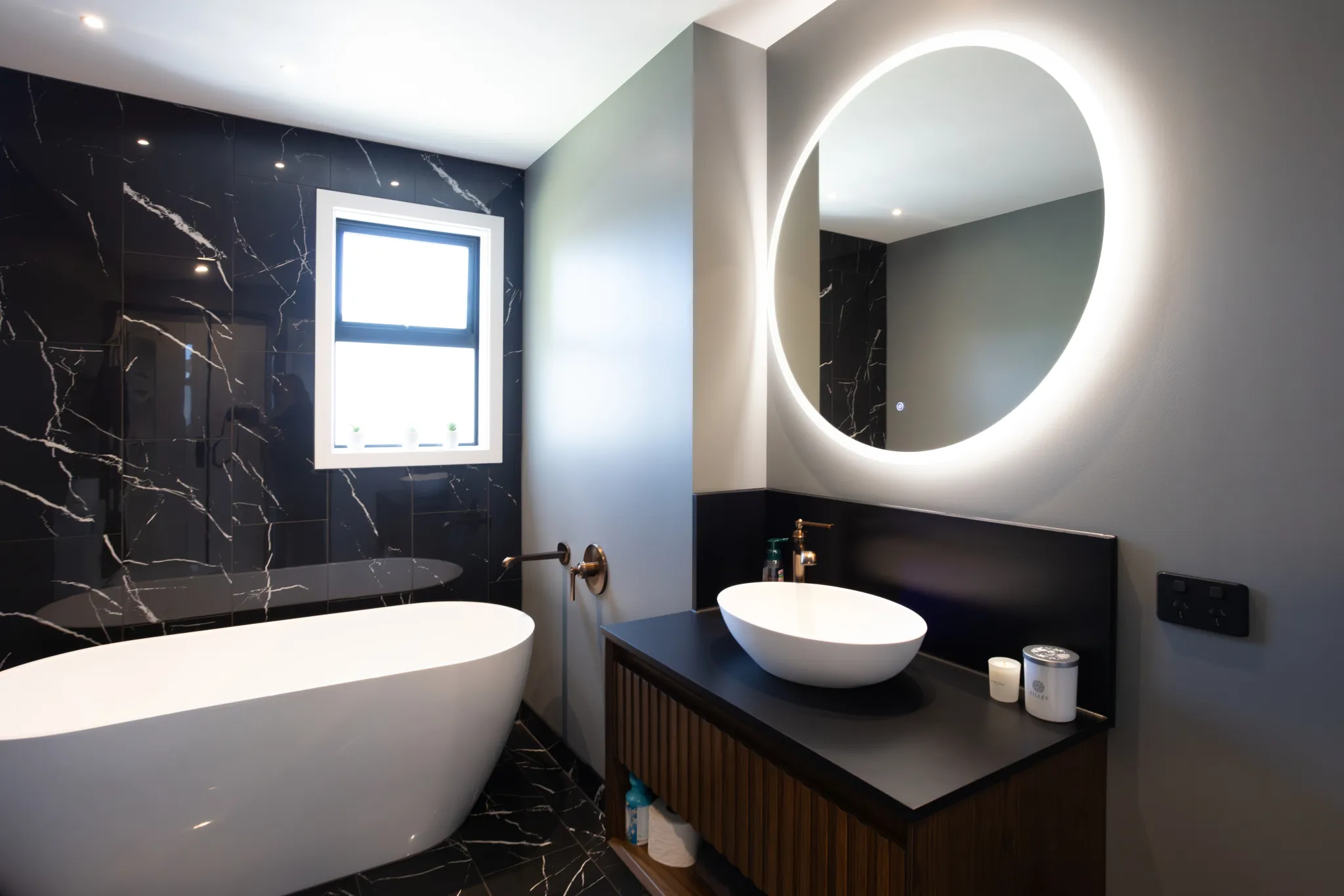 Modern bathroom with a white freestanding bathtub, black marble tiled walls, and a wall-mounted round mirror with LED backlight above a white vessel sink on a dark countertop.