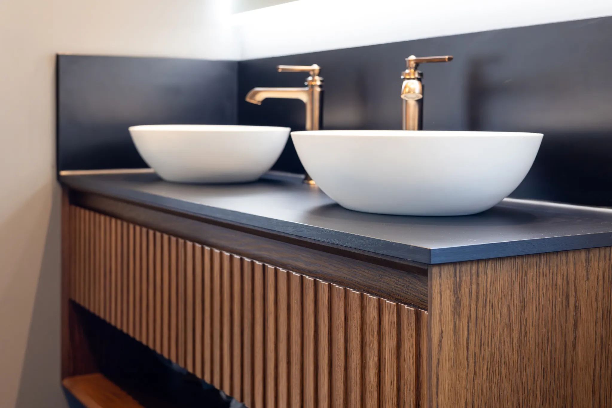 Modern bathroom vanity with two white vessel sinks, brass faucets, black countertop, and wooden ribbed cabinet.