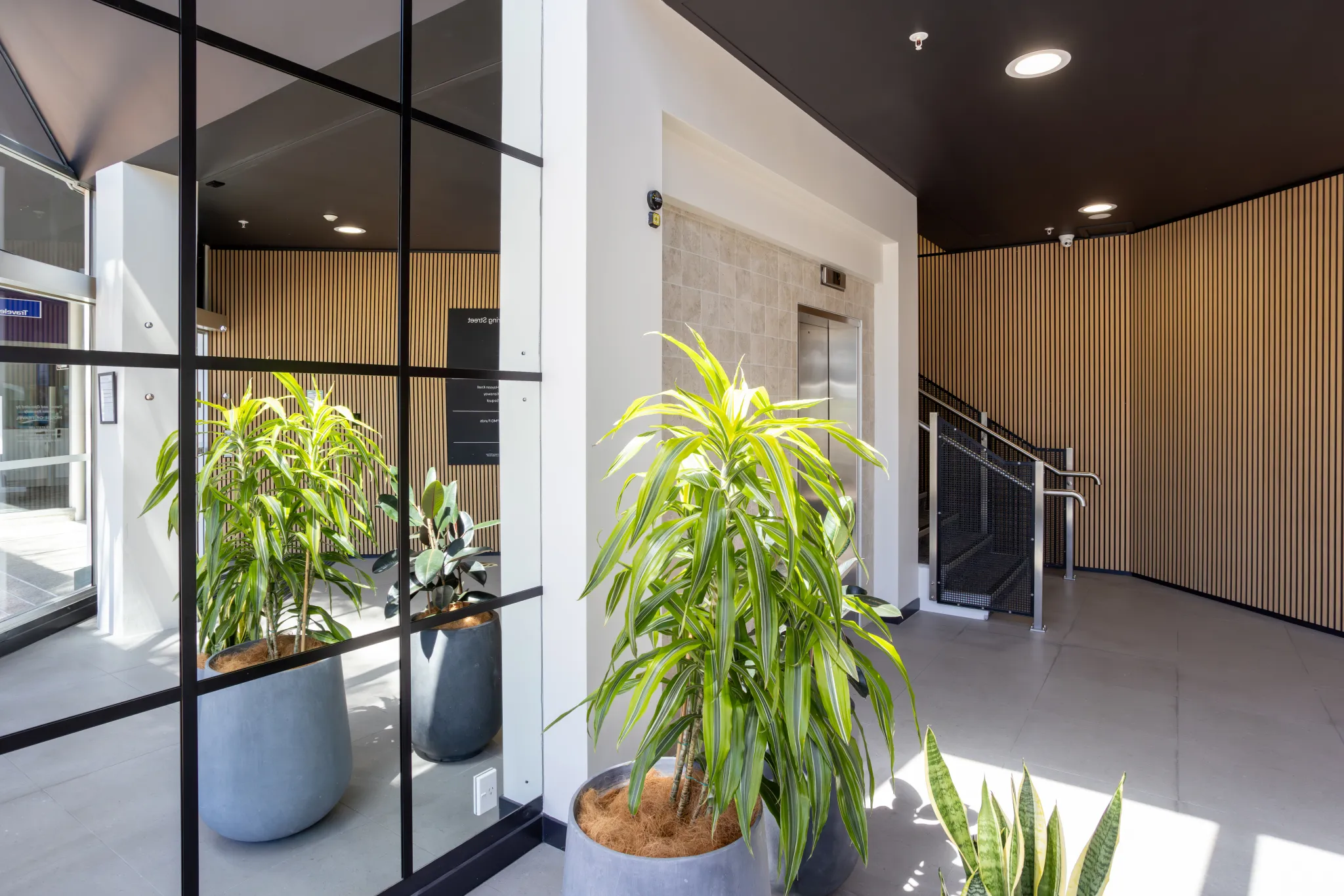 Modern building lobby with elevator, staircase, large potted plants, and vertical wood panel walls.