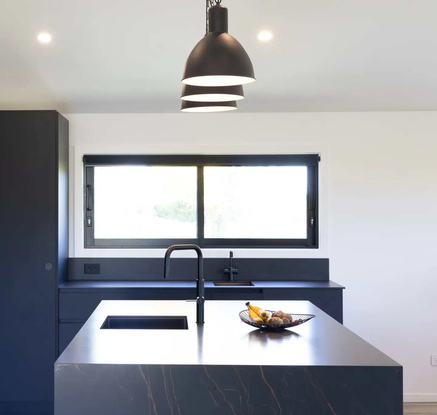 Modern kitchen with a large island featuring a black marble countertop, black sink and faucet, fruit bowl, and three hanging black pendant lights above.