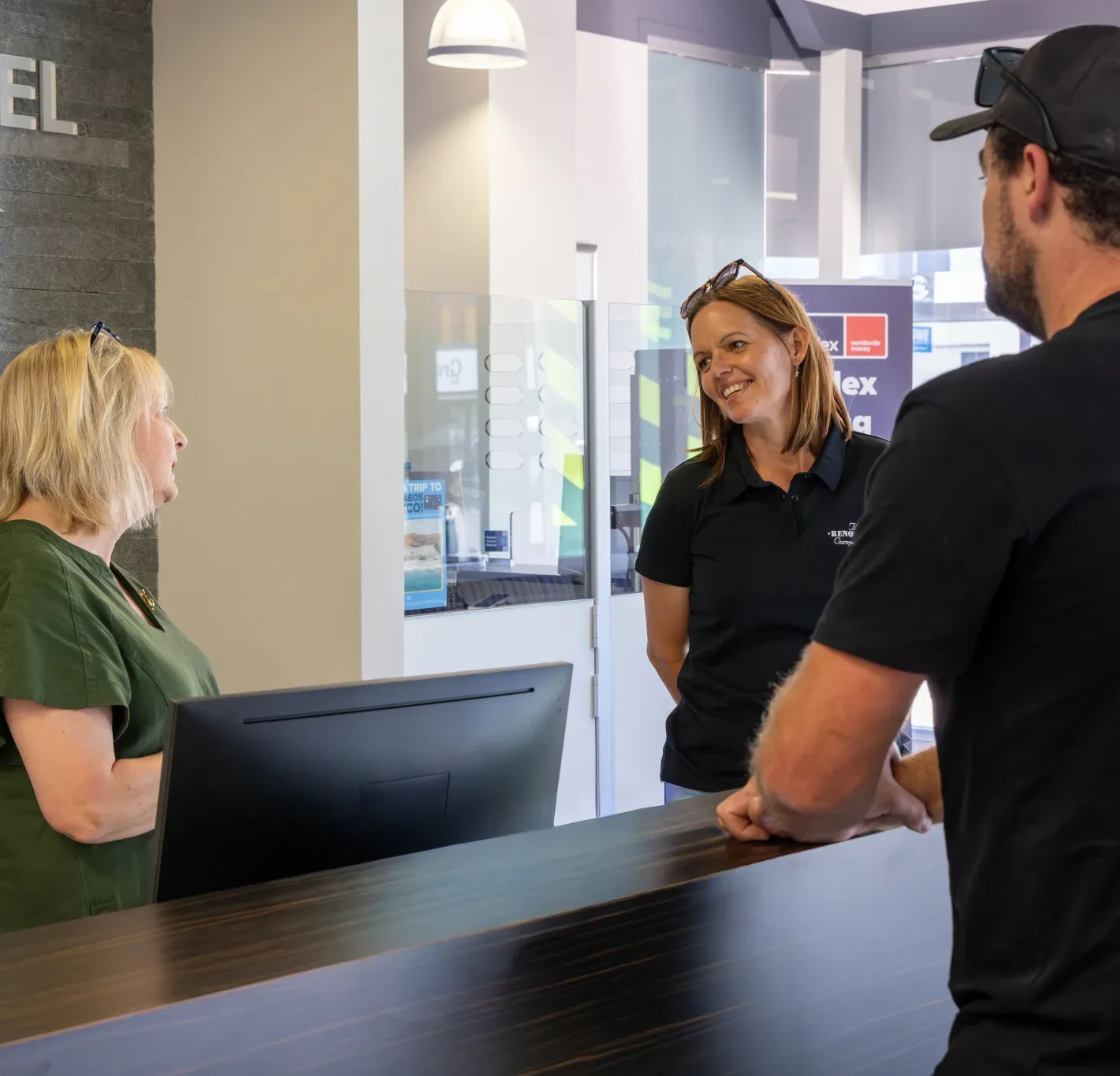 Two women and a man talking across a reception desk in a modern office with protective glass panels.