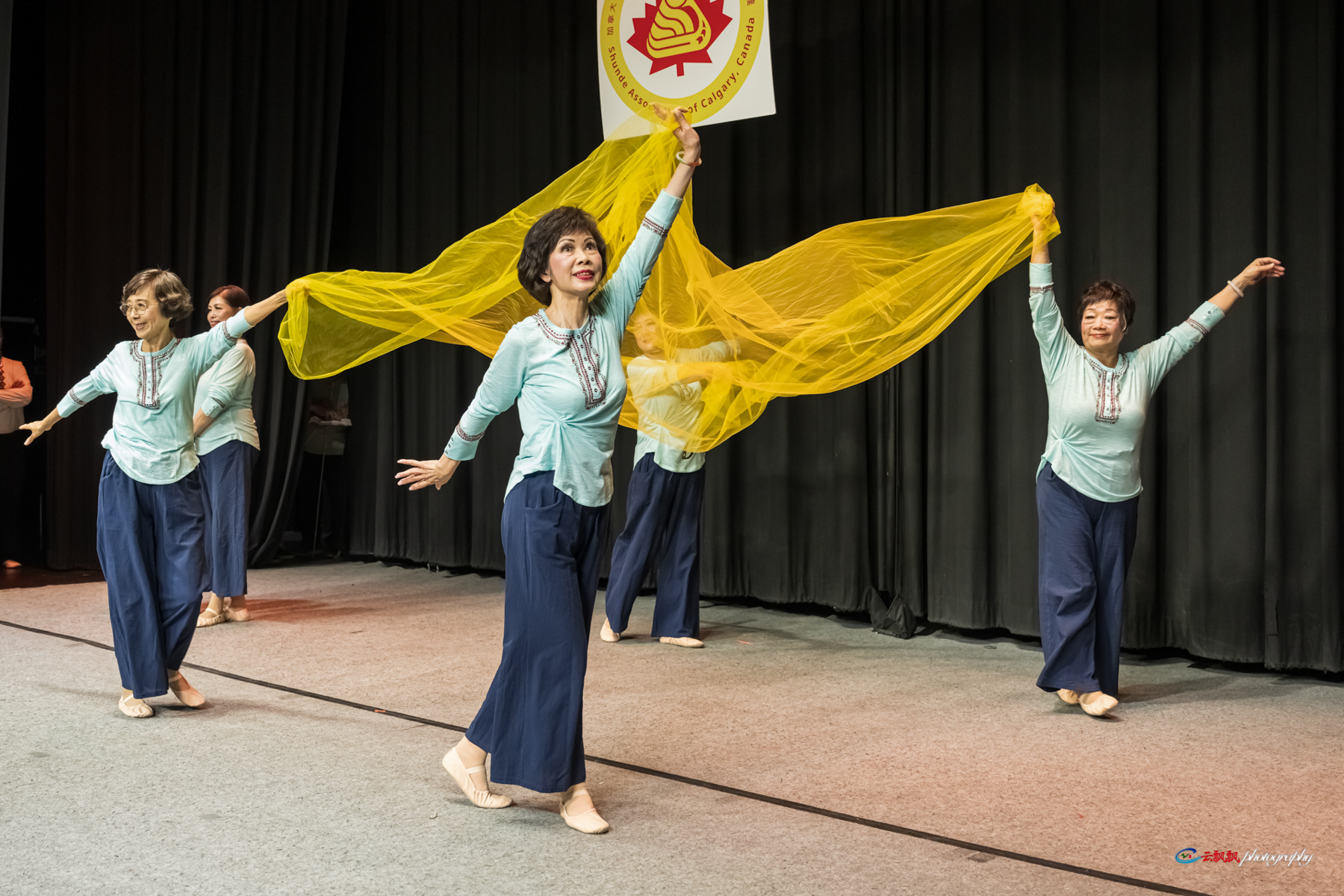 Four women in matching blue tops and pants dancing with a flowing yellow scarf on stage.