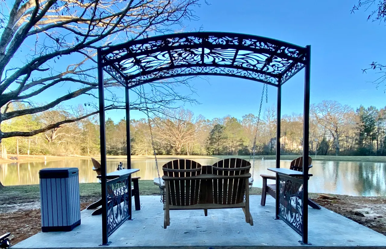 Wooden swing bench under a decorative metal trellis by a pond with trees and blue sky.