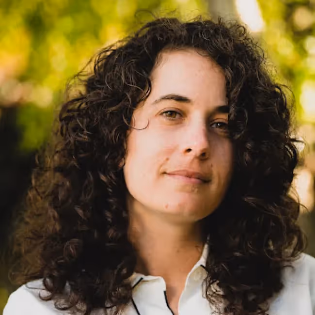 zoe glas headshot - woman with long curly dark hair in white shirt against blurred leaves in background