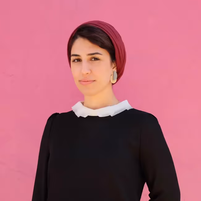 Hanieh Khosroshahi headshot - woman with short dark hair in dark shirt with red hat against a bright pink background