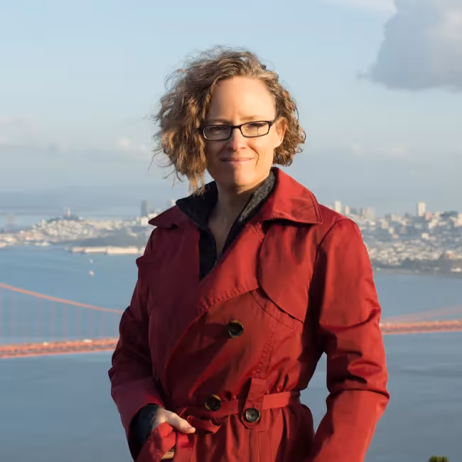 indi young headshot - woman with short reddish curly hair and glasses in a red coat standing against cityscape background