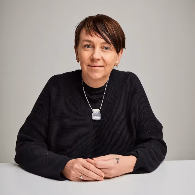 leisa reichelt headshot - woman with short dark hair in black sweater sitting at table with hands folded against white background