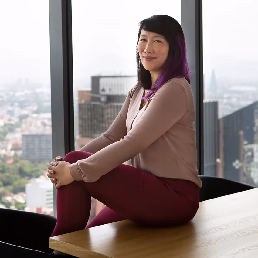 wendy johansson headshot - woman with purple hair and a pink outfit sitting on a desk in front of a window
