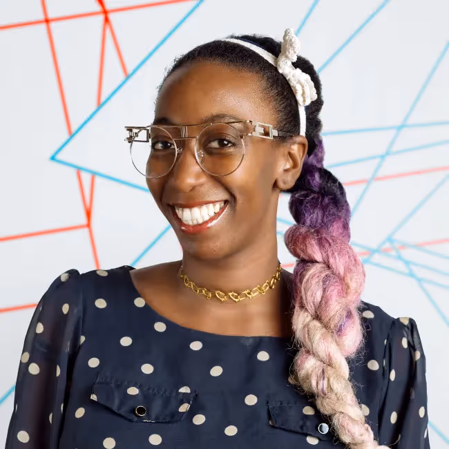 nancy douyon headshot - woman with a brown and pink braid and glasses smiling against a colorful background