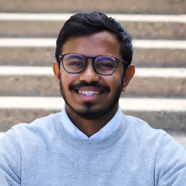 varun Murugesan headshot - man with glasses in a grey shirt standing in front of stairs