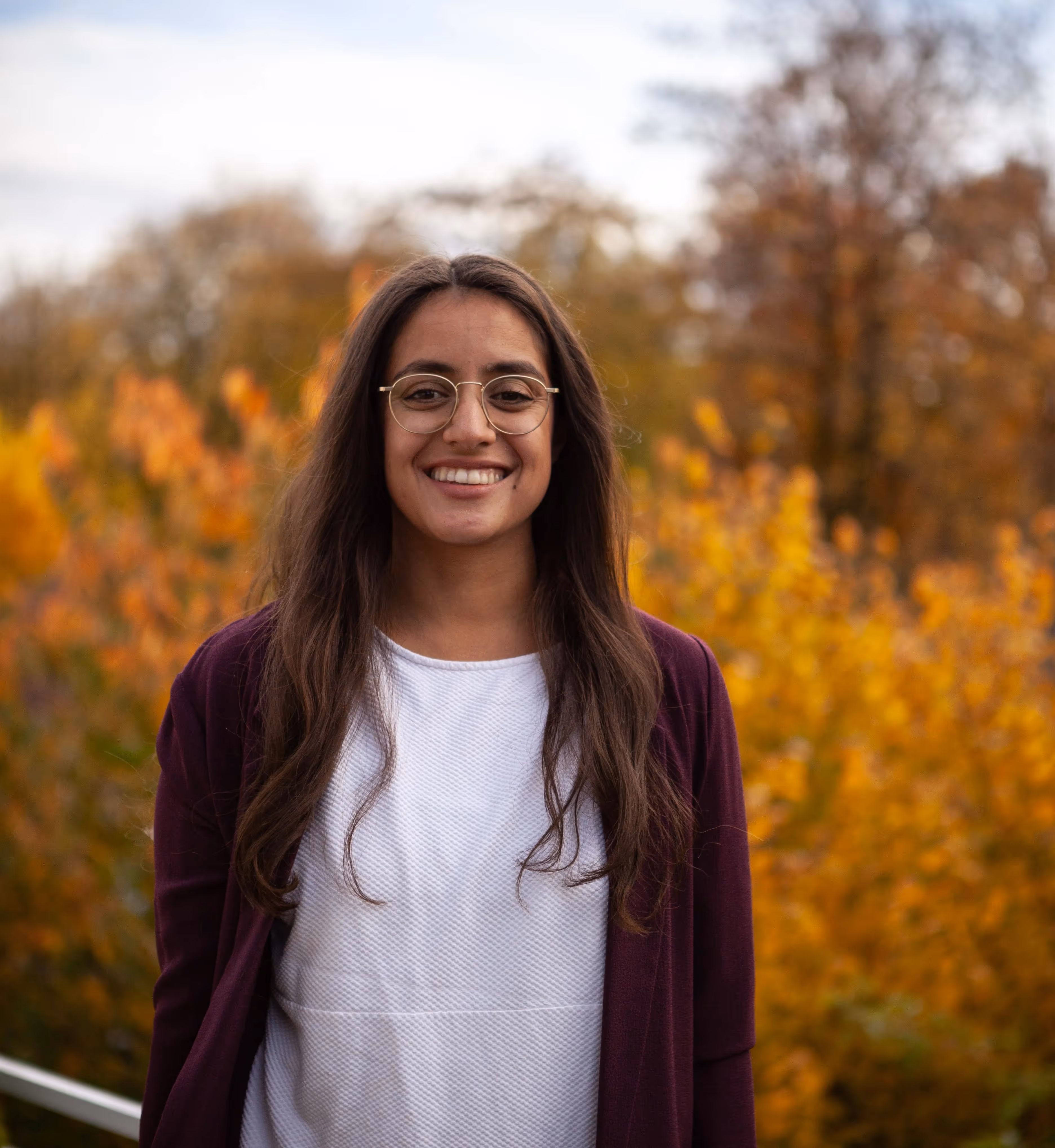 sina Schreiber headshot - woman with long dark hair and glasses against a background of yellow foliage