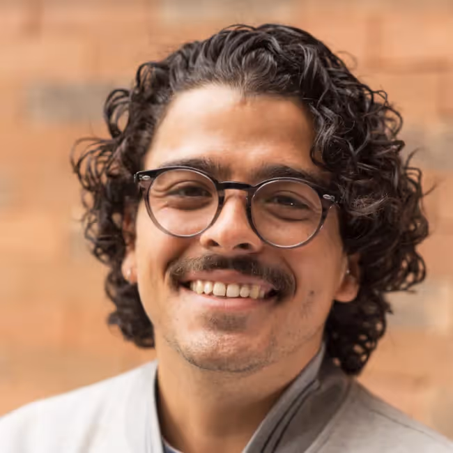 carlos hernandez tellez headshot - man with longish curly dark hair with glasses and mustache against brick background