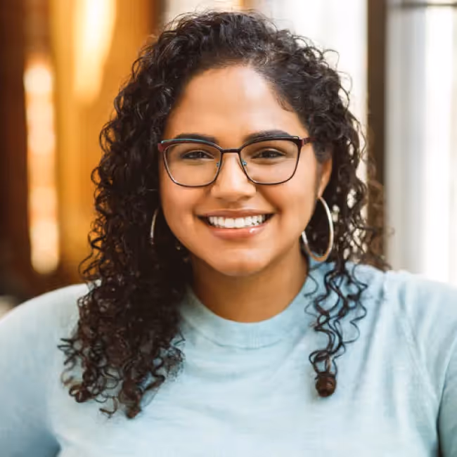 Alina I'vette Fernandez, PhD headshot - woman with long dark curly hair and black glasses in blue shirt against blurred background