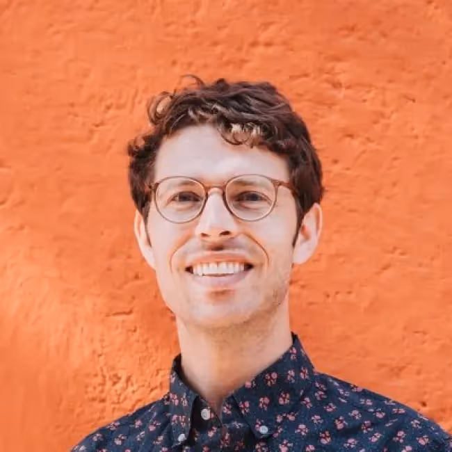 danny spitzberg headshot - man with short curly hair and glasses against an orange background
