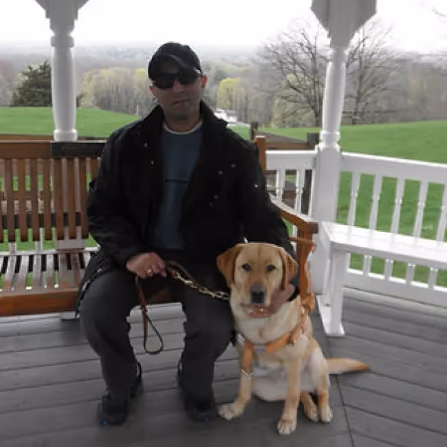 joel isaac headshot - man sitting on a bench outside with a dog against a background of trees and grass