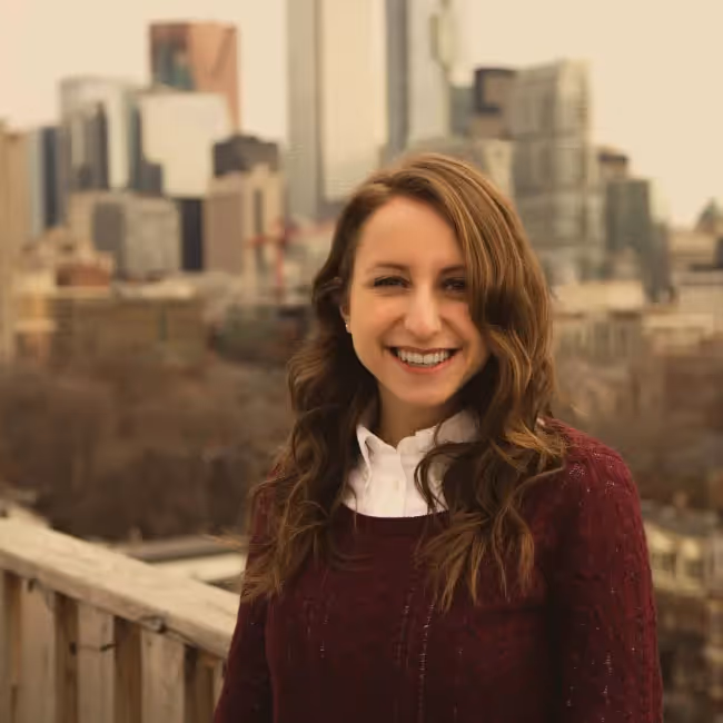 jessica baratta headshot - woman with long dark hair in maroon sweater against cityscape background