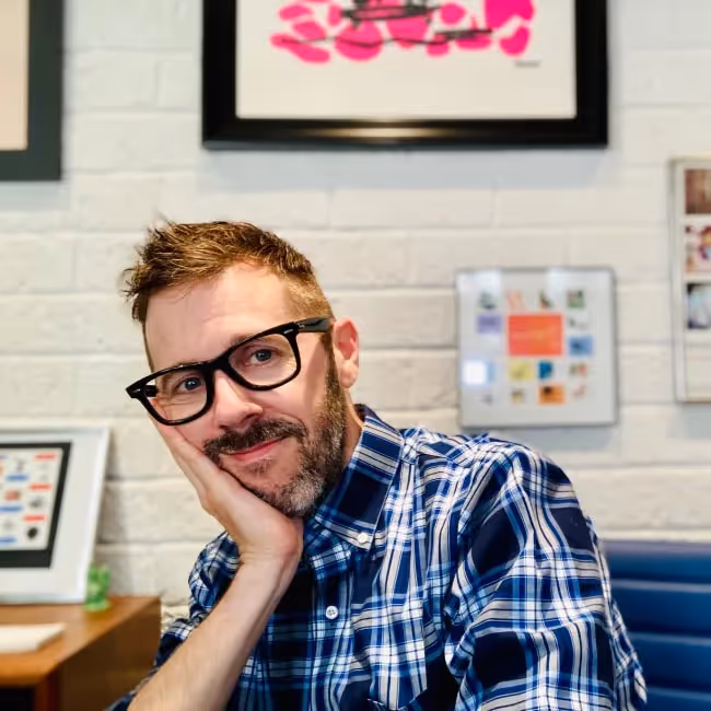 gregg bernstein headshot - man with reddish hair and beard and glasses in a blue plaid shirt leaning his head against his hand against a background of pictures on wall