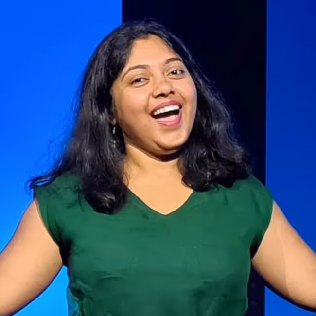 Bindu Upadhyay headshot - dark haired woman in green dress with arms held up against a blue background