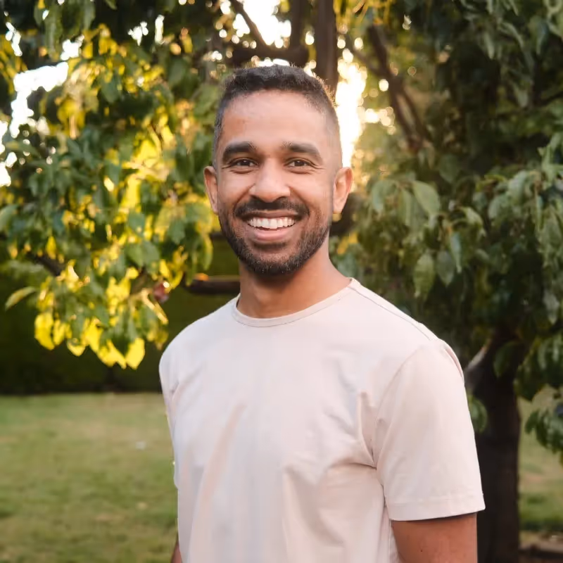 Headshot photograph of Chris Simons wearing a white shirt with a tree in the background. 