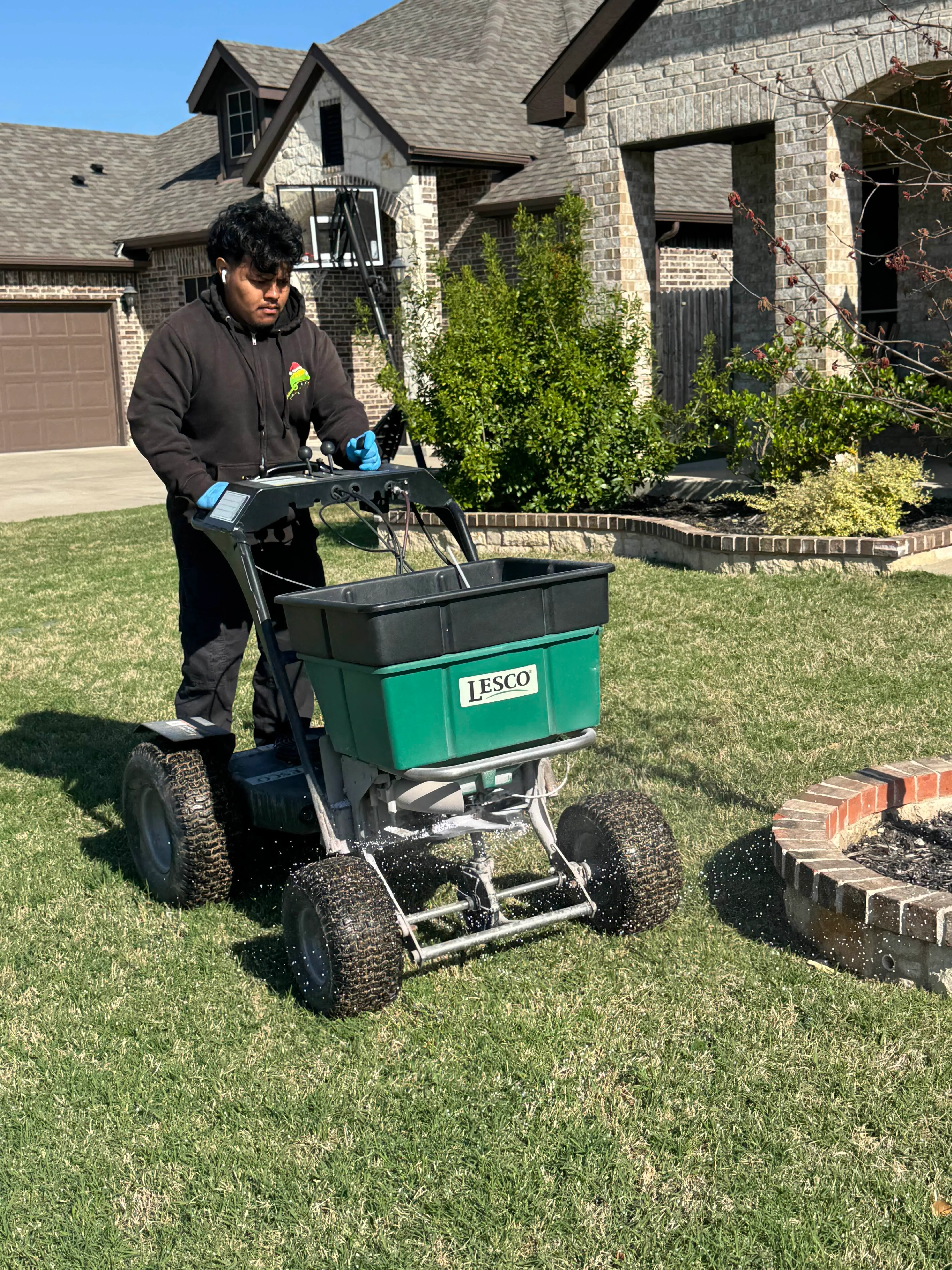 Person operating a Lesco lawn spreader on grass in front of a suburban house with stone and brick facade.