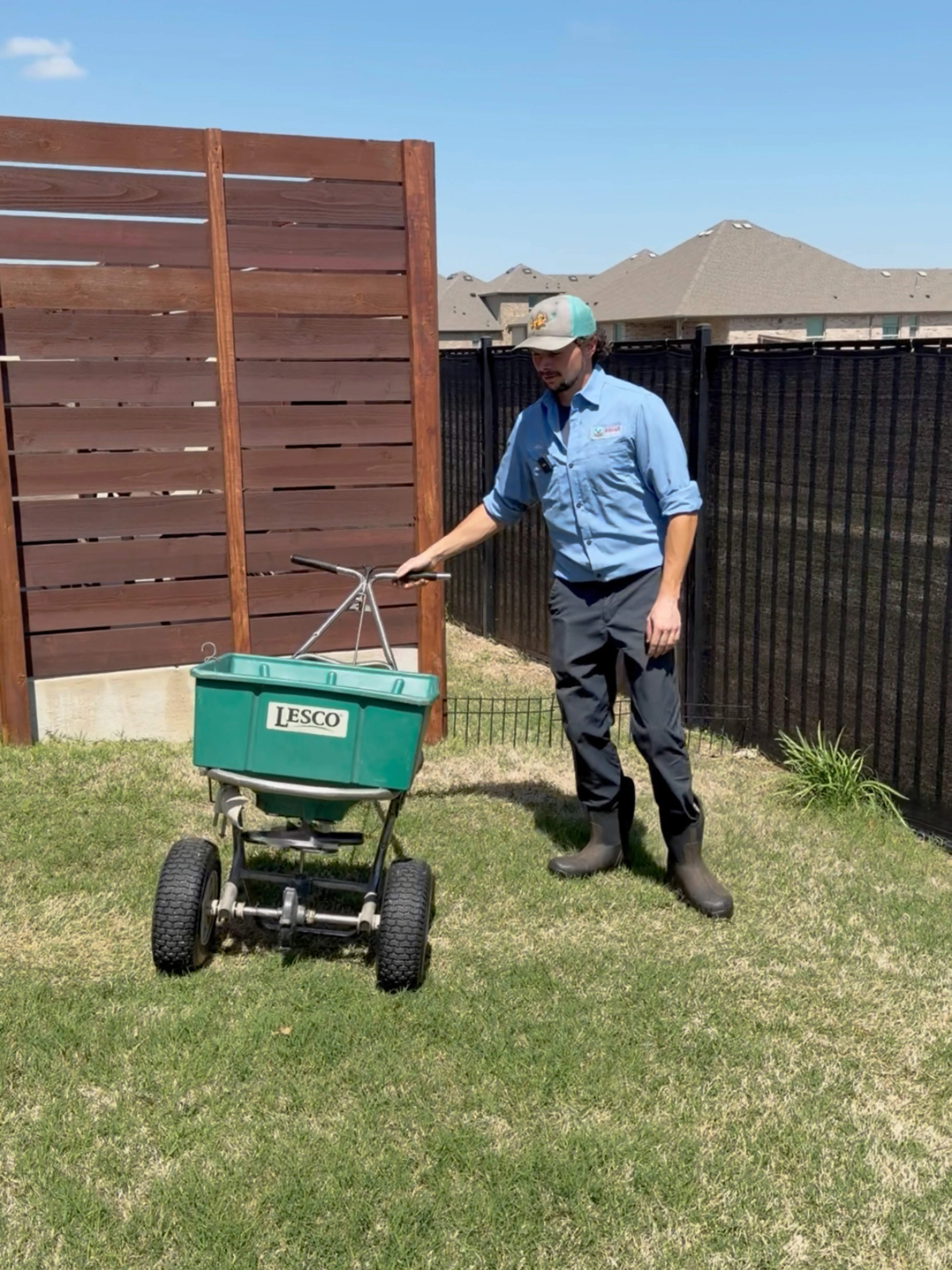 Man in uniform pushing a green Lesco lawn spreader on grass in a fenced yard.